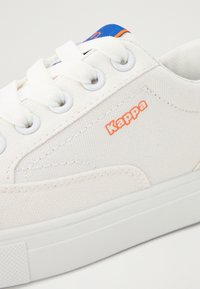 Baltos kanvasinės sportbačiai su guminiais padais, turintys oranžinį "Kappa" logotipą, baltus raiščius ir mėlyną akcentą viršuje.