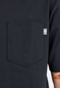 T-shirt noir à manches courtes avec une poche poitrine et une petite étiquette de marque grise. Le tissu est lisse avec une texture uniforme.