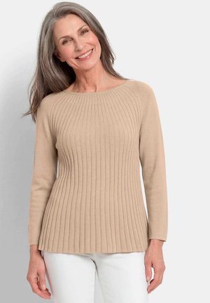 Femme d’âge moyen souriante aux cheveux gris portant un pull côtelé beige et un pantalon blanc, debout devant un fond clair uni.