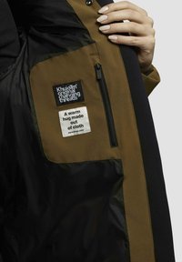 Brown and black insulated jacket interior featuring fabric tags, a zip pocket, and a label stating "A warm hug made out of cloth."