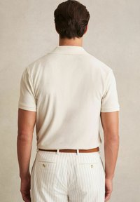 Camisa de polo de punto beige con una textura suave y mangas cortas, combinada con pantalones blancos a rayas y un cinturón de cuero marrón.
