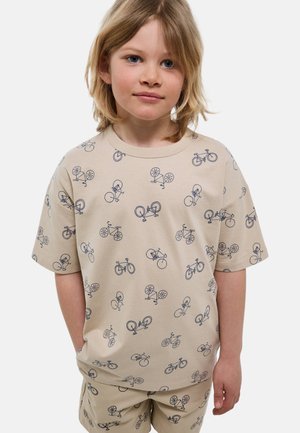 Enfant aux cheveux blonds mi-longs, portant un t-shirt beige et un short décoré d'illustrations de vélos noirs, debout les mains dans les poches.