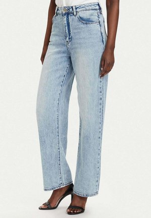 Femme portant un jean bleu clair taille haute à jambes droites et des sandales à talons noires ouvertes à l’avant, debout devant un fond blanc uni.