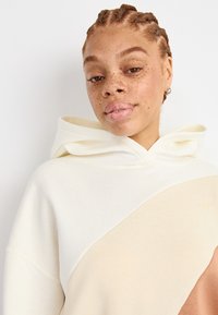 Sweat à capuche en crème et beige avec un design color-block. Tissu doux, coupe décontractée, avec une grande capuche et des détails de couture visibles.
