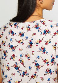 Topp med kort ärm i en ljusbeige tyg med ett blommönster i blått, rött och gult. Slät textur med rundad halsringning.