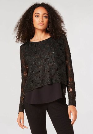 Top nero all'uncinetto con una vestibilità ampia, design a rete intricato e maniche lunghe. Sotto c'è una blusa nera liscia.