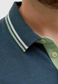 Marineblauw poloshirt van een gestructureerde stof, met een groene kraag met witte strepen en twee knopen, dat het unieke ontwerp benadrukt.