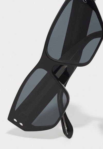 Lunettes de soleil noires surdimensionnées avec des verres sombres, cadre angulaire et matériau léger. Présente des accents matériels subtils sur le cadre.