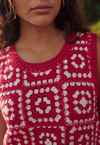 Gehaakte top met een geometrisch patroon in rood en wit. Het ontwerp bevat vierkanten en ruiten met gegolfde randen en een ronde halslijn.