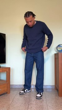 Suéter de malha texturizada azul-marinho, jeans retos azuis e ténis preto/e branco. Em pé sobre um chão de granito claro com móveis de madeira nas proximidades.