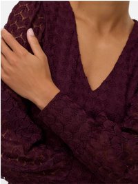 Nærbilde av kryssete armer iført en dyp lilla teksturert bluse i blonder med V-hals.