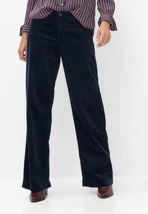 Personne portant un pantalon large en velours côtelé bleu foncé, des chaussures marron à talons, et une chemise rayée marron et blanche à manches longues.