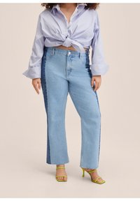Personne aux formes portant un jean droit bicolore bleu clair et bleu foncé, une chemise rayée nouée et des sandales à talons vert lime sur fond beige.