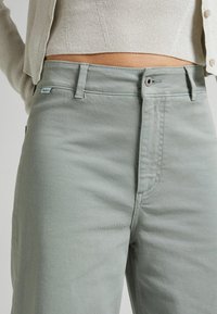 Vaaleanvihreät puuvillasekoitetekstiilisestä valmistetut housut suoran leikkauksen kanssa, joissa on etutasku ja metalliset detaljit. Sileä pinta, minimaalinen ompelu.