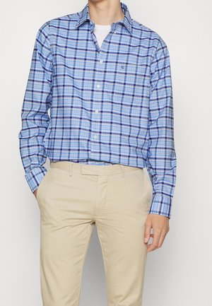 Homme portant une chemise à carreaux bleue et blanche boutonnée sur un t-shirt blanc, avec un pantalon beige, debout devant un fond clair uni.