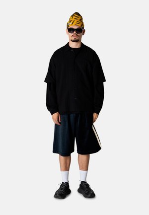 Mand står iført sort skjorte med knapper, mørke shorts med sideliste, sorte sneakers, hvide sokker, gul mønstret hat og store sorte solbriller.