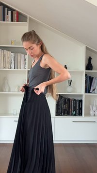 Vrouw met lang haar in een grijs tanktop die een zwarte plooirok aantrekt in een kamer met witte planken en boeken.