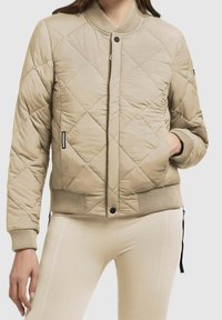 Beżowa pikowana kurtka z prążkowanym kołnierzem i mankietami, wyposażona w zapięcie na zatrzaski oraz boczne kieszenie na zamek. Gładka faktura materiału.