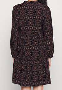 Svart klänning med ett diamantramönster i rött och vitt, långa ärmar, figurnära midja och ett flerlagerskjoldesign.