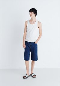 Débardeur blanc sans manches, short en denim bleu à mi-cuisse et sandales noires avec des accents blancs sur un modèle devant un fond uni.