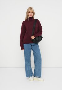 Pulover cu guler înalt de culoare burgundă, asortat cu blugi evazați de culoare albastru deschis și adidași albi. O geantă crossbody moale, de culoare neagră, completează ținuta.