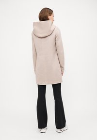 Beiger Wollmantel mit großer Kapuze, geradem Schnitt und minimalen Nähten, kombiniert mit schwarzen Schlaghosen und weißen Sneakers.