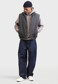 Junger Mann, der nach unten schaut, trägt eine gemusterte Steppweste über einem grauen Hoodie, lockere dunkelblaue Jeans, weiße Sneaker und eine marineblaue Mütze.