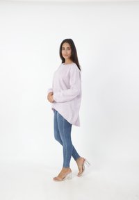 Lila oversized trui met een ronde hals en verlengde schouderlijnen, gecombineerd met strakke blauwe jeans en doorzichtige hak sandalen op een witte achtergrond.