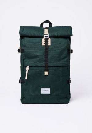 Dark green roll-top backpack with black strap and silver buckle, beige zipper pull, side buckles, and white label reading Sandqvist.