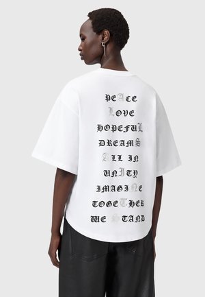 Hvid oversized T-shirt med sort og sølv tekst trykt lodret på ryggen. Rund hals og korte ærmer, lavet af bomuldsstof.