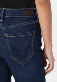 Tmavomodré džínsy z denimu s kontrastným šitím, so výrazným dizajnom zadnej vrecka a hnedou koženou značkovou nálepkou na páse.