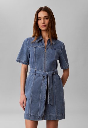 Mujer que lleva un vestido de mezclilla azul claro de manga corta con cremallera frontal y lazo en la cintura, de pie frente a un fondo gris liso.