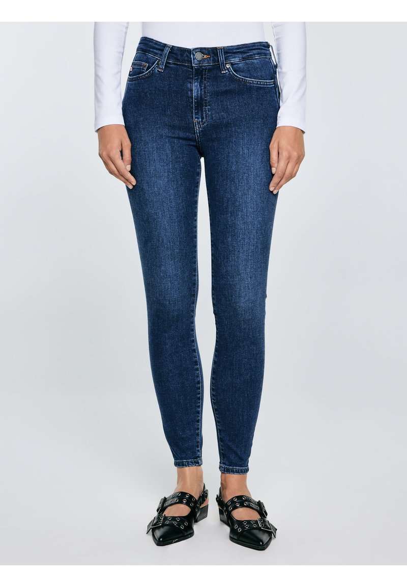 Naine, kes kannab tumelisi siniseid kitsaid teksaseid, valget pika varrukaga särki ning musti terava ninaga baleriinikingi konksude ja silmustega.