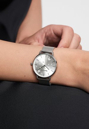 Silver wristwatch with star-patterned dial, metal mesh strap, round case, and simple hour markers. Positioned on a neutral background.