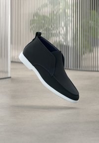 Chaussure noire en cuir à enfiler, avec une surface lisse, une semelle blanche et des coutures bleues contrastantes. Présente un profil bas et un design minimaliste.