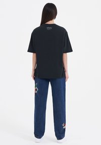 Black oversized t-shirt with short sleeves and a high neckline, worn with loose-fitting blue denim pants featuring colorful embroidery.