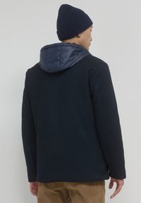 Giacca esterna blu navy con un corpo in maglia testurizzata e un cappuccio liscio e gonfio. Completata con un berretto a coste e indossata sopra pantaloni color khaki.