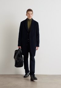 Schwarzer Wollmantel über einem grünen Rollkragenpullover, kombiniert mit dunklen Hosen und schwarzen Schuhen. Trägt eine schwarze Sporttasche mit einem runden Akzent.