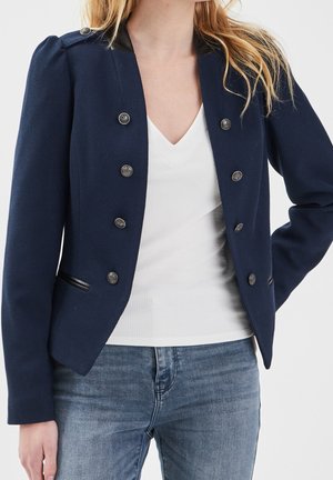 Femme portant un blazer bleu marine croisé à boutons argentés, un haut blanc à col en V et un jean bleu devant un fond uni.