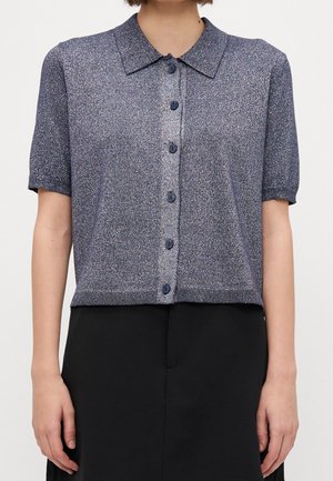 Vrouw draagt een kort mouw, knoopjes, glanzend gestructureerd blauw vest in combinatie met een zwarte rok tegen een effen achtergrond.