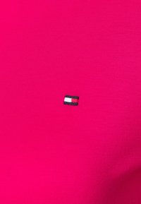 Lys pink bomuldsstof med et lille broderet logo i rød, hvid og sort placeret nederst til venstre. Glat tekstur.