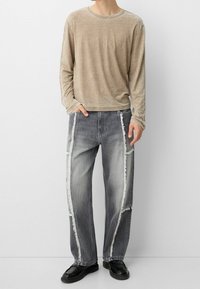 Béžové tričko s dlouhým rukávem spojené se šedými džínovými kalhotami s roztřepenými bočními švy. Boty modelu jsou černé a kalhoty mají volný střih.