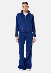 Blauer Trainingsanzug aus weichem Stoff. Zip-Jacke mit hohem Kragen und drei weißen Streifen an den Ärmeln. Weit geschnittene Hose mit passenden Streifen.