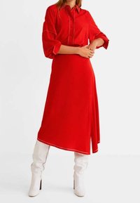 Robe rouge à boutons avec une coupe ample, dotée de manches trois-quarts, d'une fente sur le côté et d'accents de couture blanche. Associée à des bottes blanches jusqu'aux genoux.