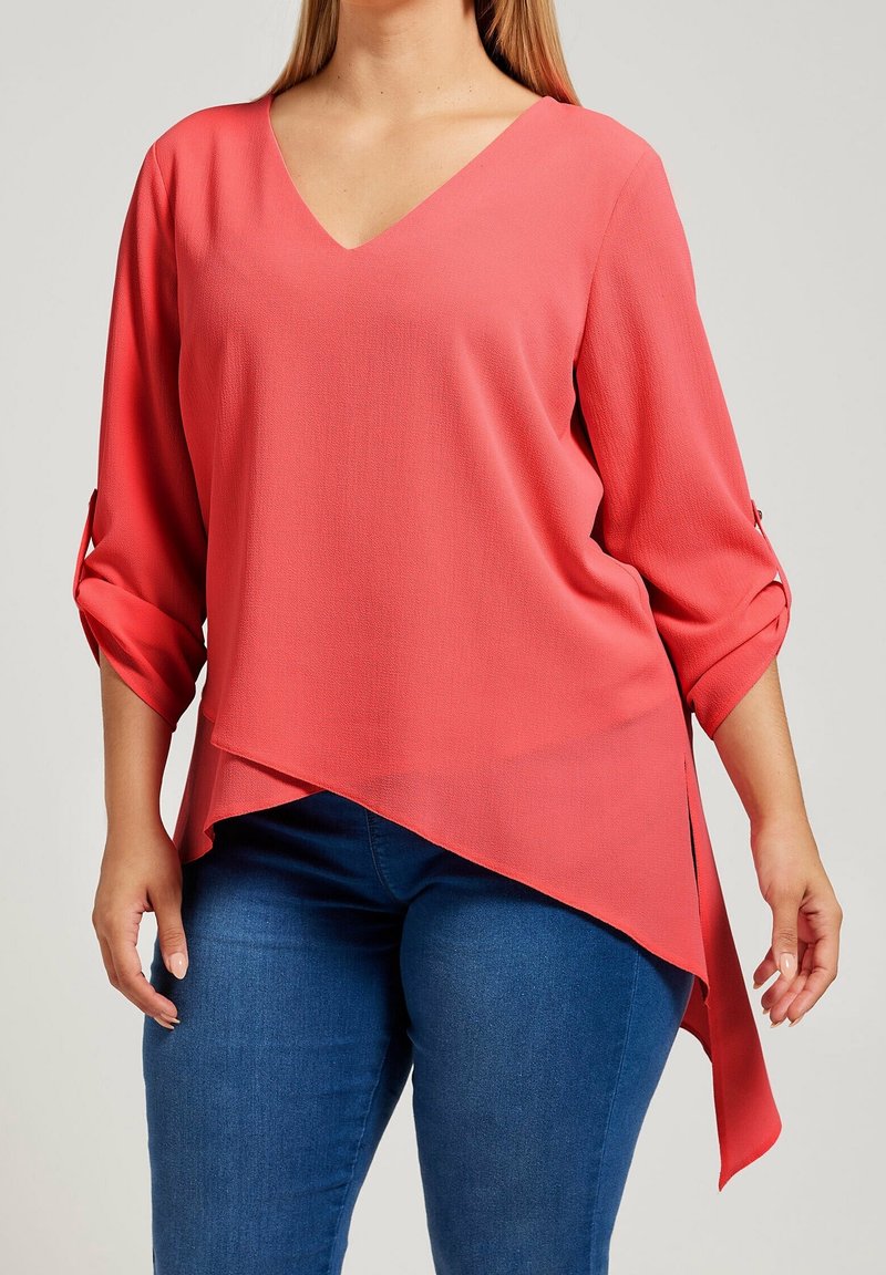 Top corallo con scollatura a V, maniche a tre quarti, orlo asimmetrico e polsini arrotolati. Realizzato in tessuto morbido e leggero. Abbinato a jeans blu.