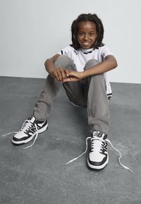 Baskets noires et blanches avec lacets, en cuir texturé, bout rond, arborant le logo New Balance. Portées avec un jean gris et un t-shirt graphique.