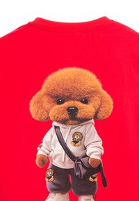 Camiseta roja con un gráfico de un personaje de perro de peluche que lleva una sudadera con capucha blanca y pantalones anchos negros, con un diseño de oso bordado.
