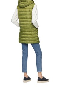 Olivgrüne Pufferweste mit Kapuze, die über gesteppten Bereichen verfügt. Getragen über einem weißen Oberteil, kombiniert mit blauen Jeans und dunklen Schuhen.