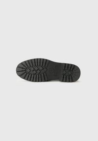 Schwarze Gummischuhsohle mit einem strukturierten Grip-Muster, tiefen Rillen und einem zentralen Logo-Abdruck. Flaches und breites Design.