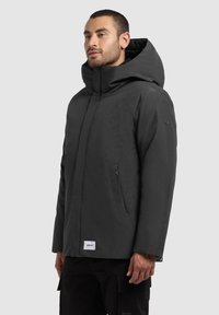 Dark gray hooded jacket with a zippered pocket, minimalist design, and a logo patch on the hem. Made from a smooth, water-resistant material.